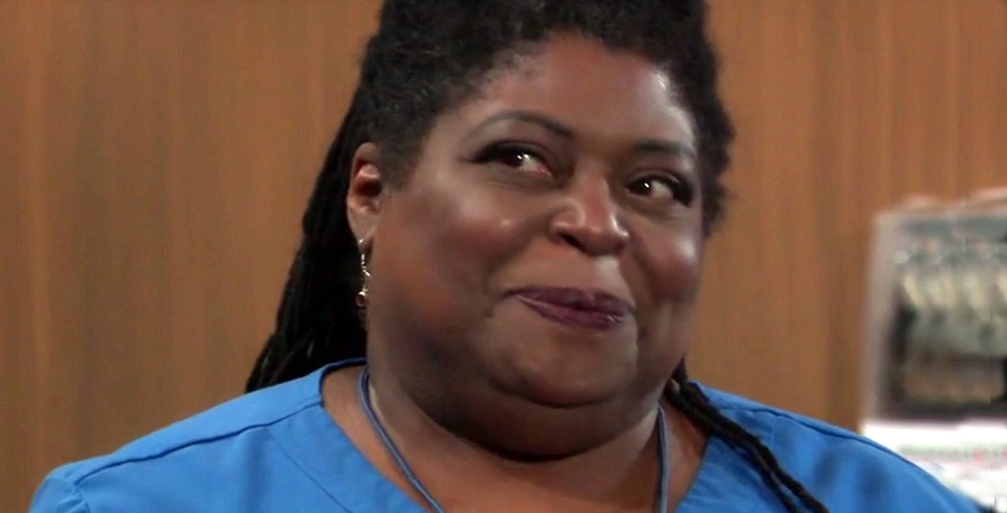 sonya eddy as nurse epiphany johnson general hospital wearing blue scrubs in front of a brown wall