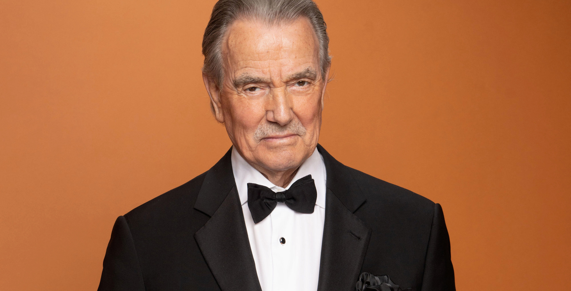 eric braeden of the young and the restless against an orange background in a tux